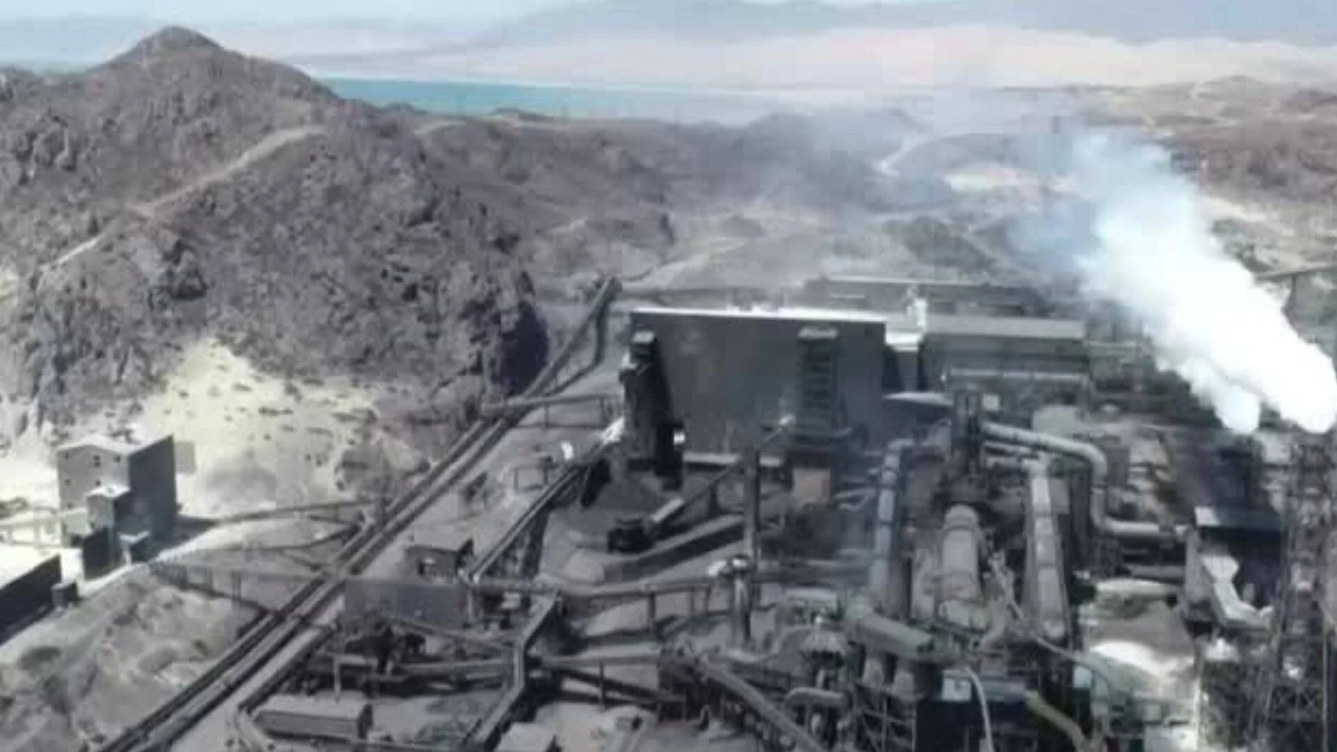 Vista aérea de una gran planta industrial con emisiones de humo blanco, rodeada de montañas áridas y un lago al fondo.