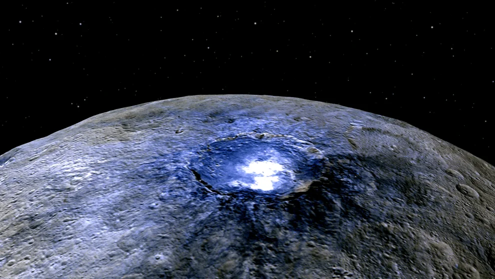 Vista orbital de un cráter con manchas blancas luminosas sobre la superficie gris de un cuerpo celeste rocoso y el espacio estrellado al fondo.