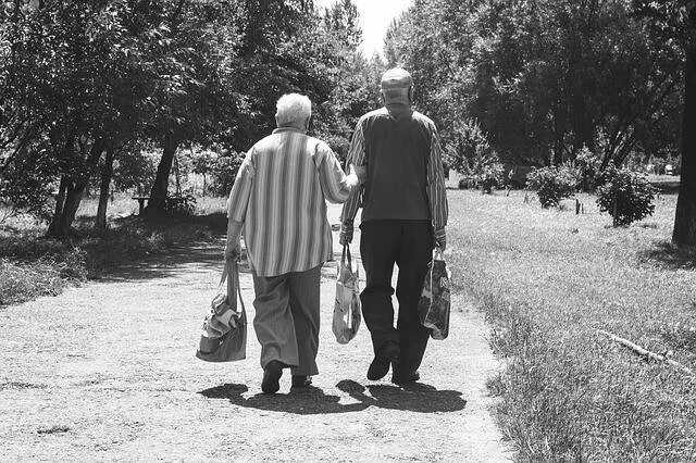 Dos personas mayores caminan de espaldas por un camino rodeado de árboles, llevando bolsas de la mano.