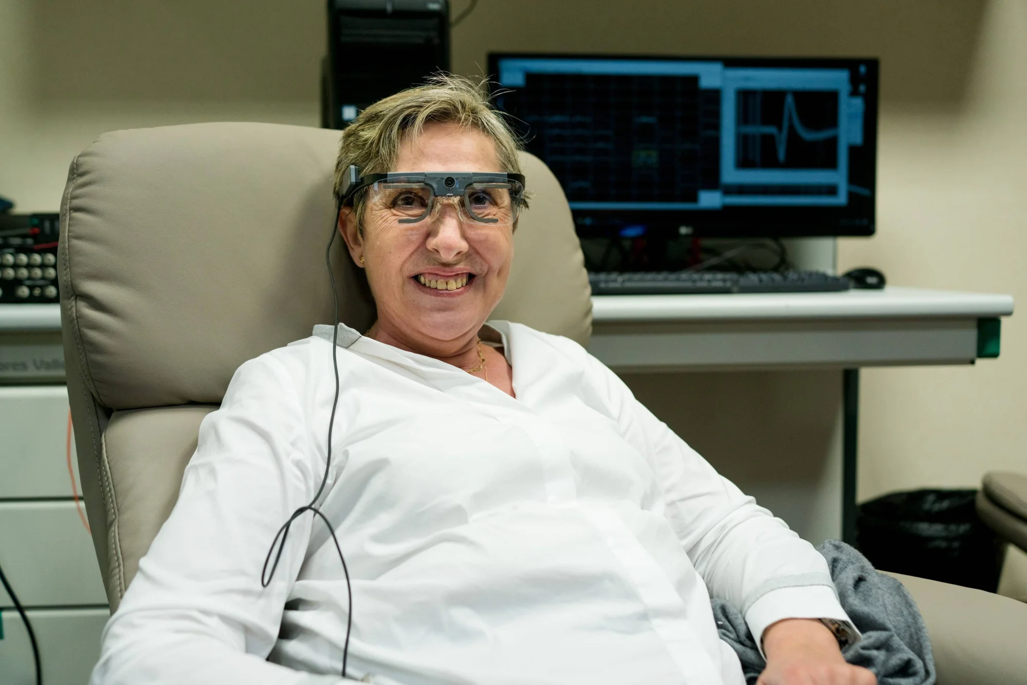 Mujer mayor sonriente recostada en sillón clínico, con gafas de tecnología visual conectadas y monitor médico al fondo.