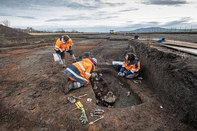 Tres arqueólogos con chalecos naranjas trabajan en una excavación arqueológica, examinando restos y objetos en el terreno.