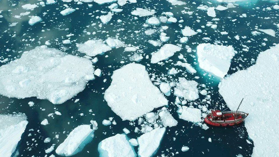 Vista aérea de un pequeño barco rojo navegando entre témpanos de hielo flotantes en aguas árticas.