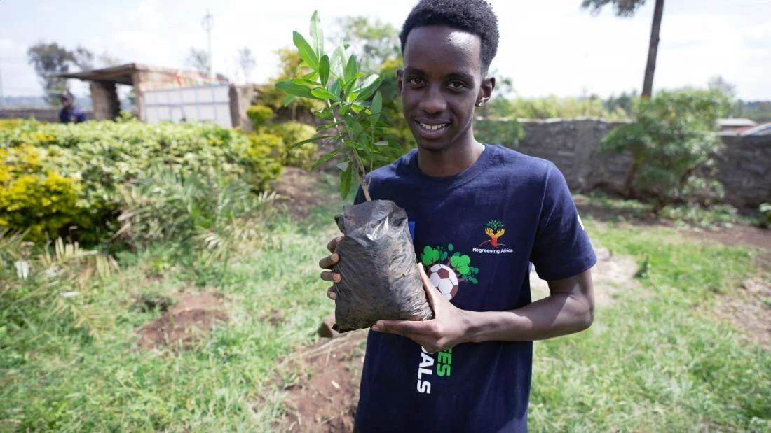 Joven sonriente con camiseta de Regreening Africa sostiene una plántula lista para plantar en un área verde.