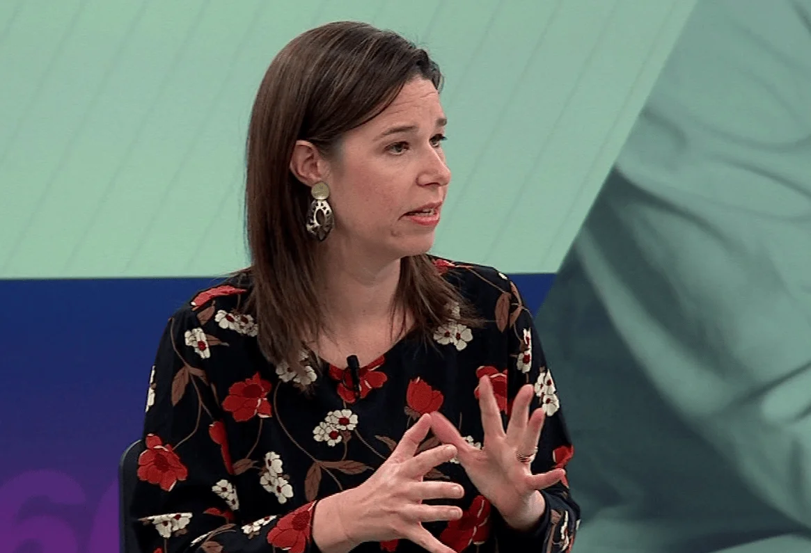 Mujer con blusa floral negra gesticula con las manos mientras habla en un estudio de televisión con fondo verde.