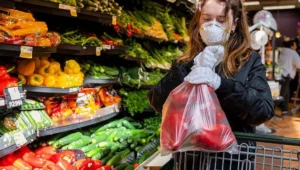 Mujer con mascarilla FFP2 y guantes blancos selecciona verduras en la sección de frescos de un supermercado.