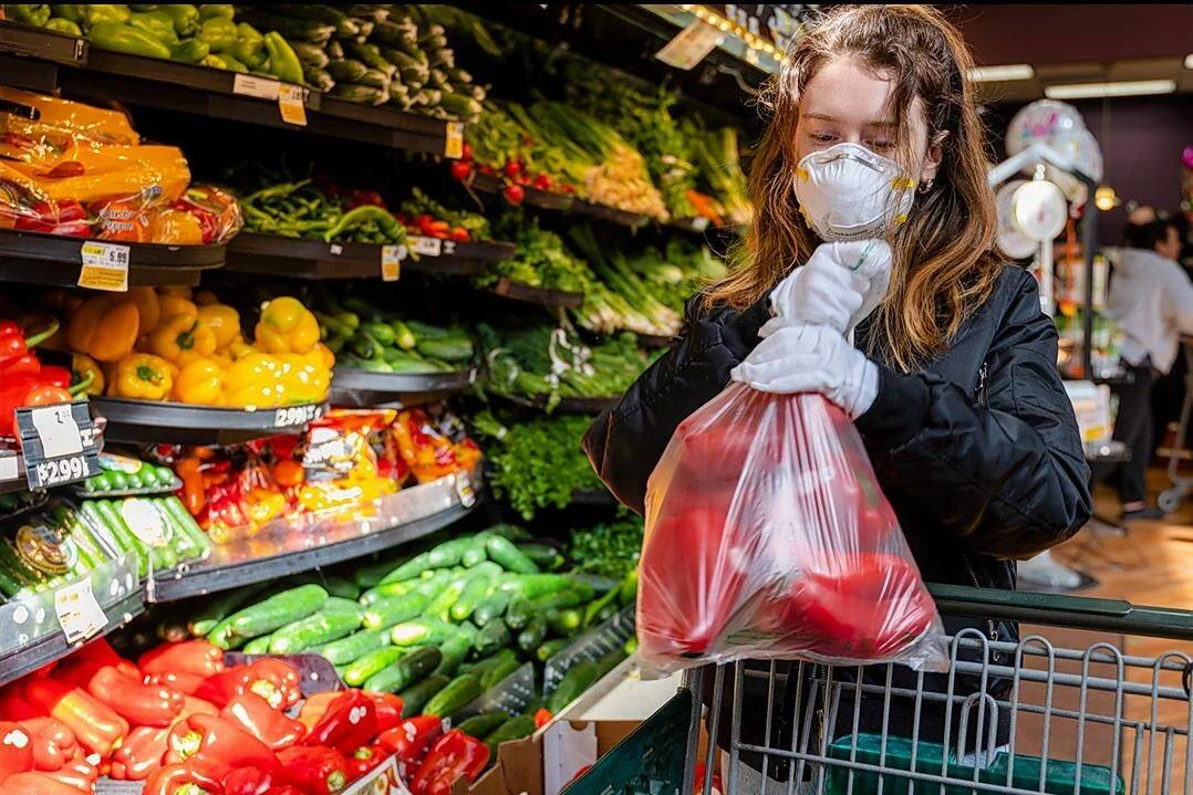 Mujer con mascarilla FFP2 y guantes blancos selecciona verduras en la sección de frescos de un supermercado.
