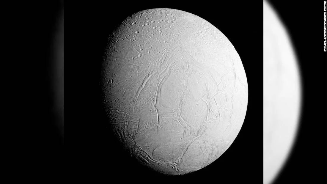 Vista en blanco y negro de Encélado, luna de Saturno, mostrando su superficie helada con grietas y cráteres captada por la sonda Cassini.