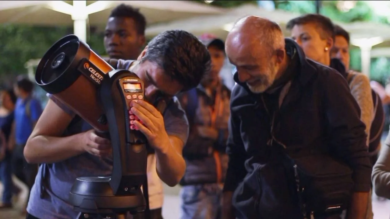Un hombre mira a través de un telescopio Celestron NexStar rodeado de personas en una actividad de astronomía nocturna al aire libre.