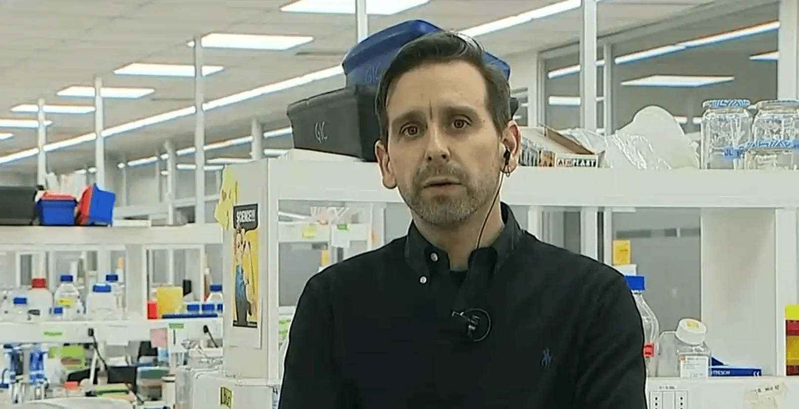Hombre con micrófono de solapa habla frente a cámara en un laboratorio equipado con estantes y material científico.