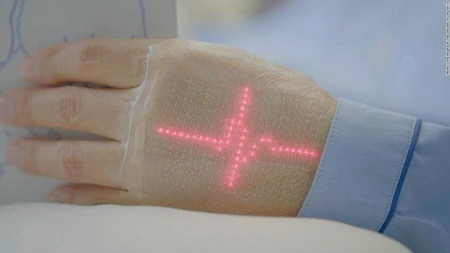 Parche electrónico translúcido pegado en el dorso de una mano que muestra una cruz luminosa de puntos LED rosados.