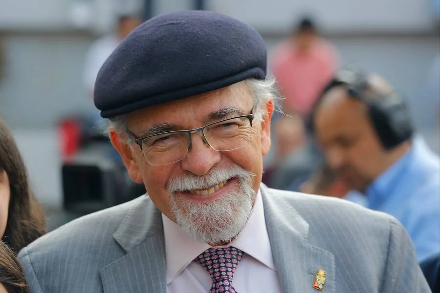 Hombre mayor con barba blanca, boina negra, gafas y traje gris sonríe en un evento público.