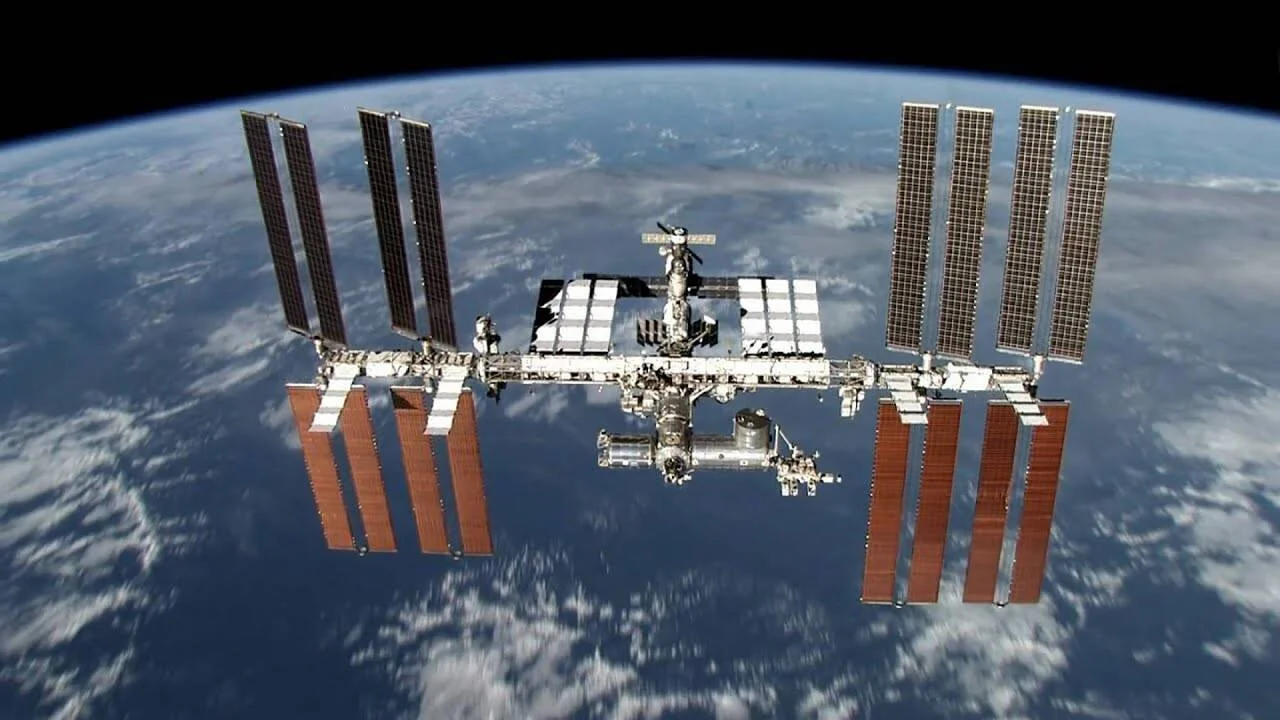 La Estación Espacial Internacional con sus paneles solares desplegados orbita sobre la Tierra con nubes visibles al fondo.