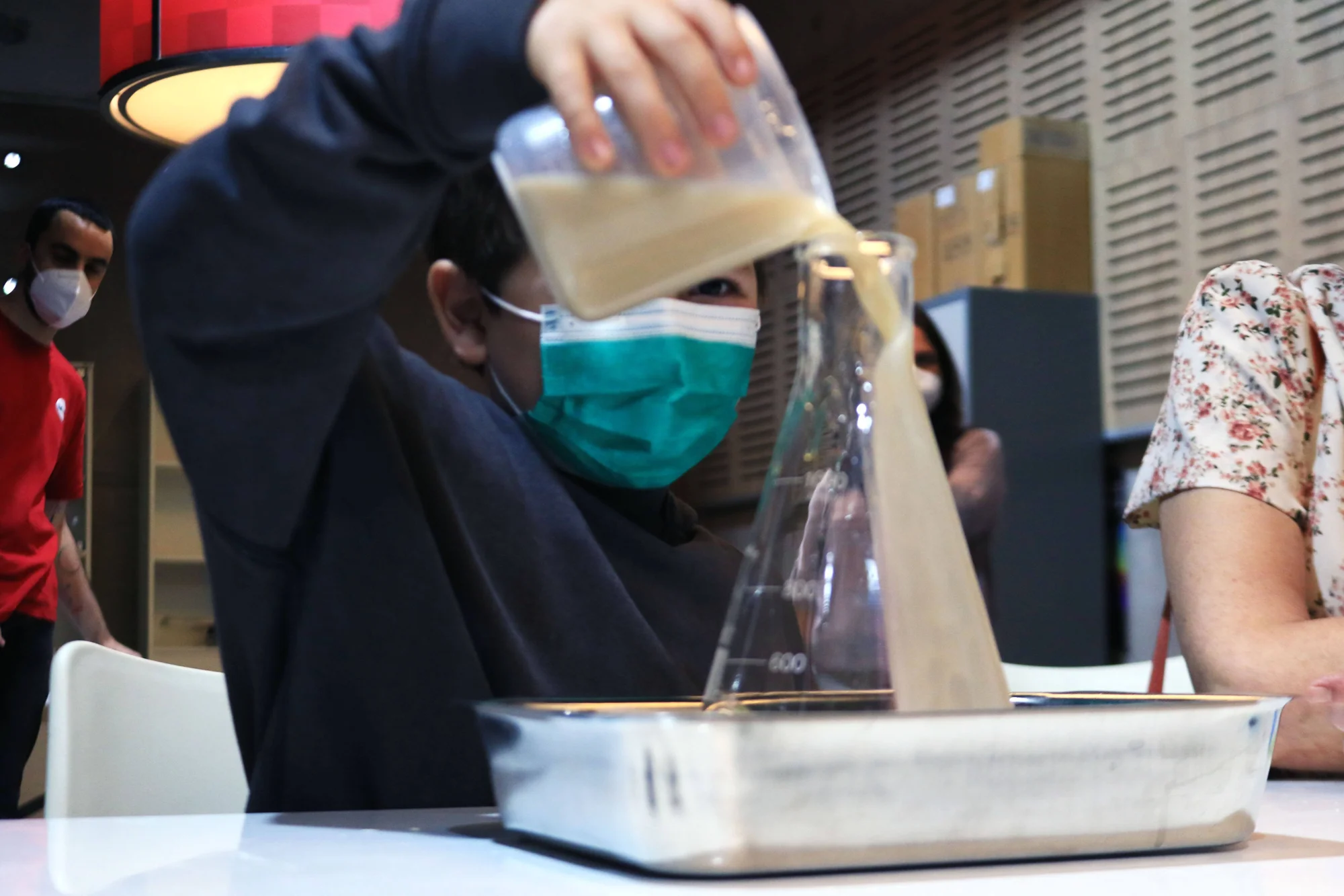Un niño con mascarilla vierte una sustancia marrón desde un recipiente plástico hacia un matraz de laboratorio durante una actividad educativa.