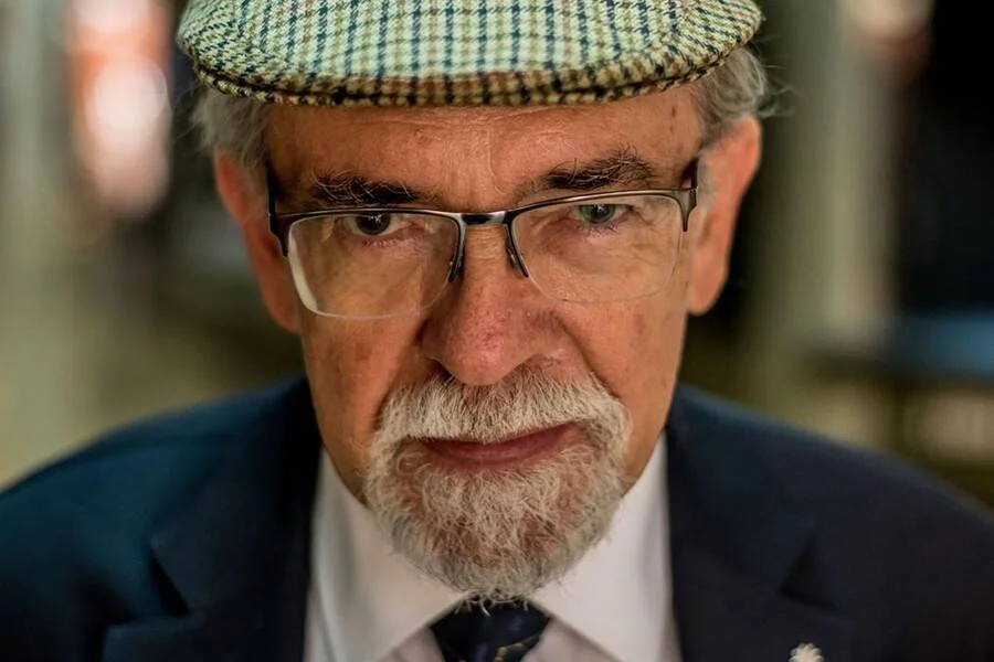 Hombre mayor con barba blanca, anteojos, gorra de cuadros y traje azul marino mira fijamente a la cámara.