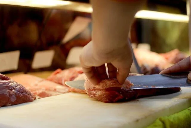 Manos de un carnicero cortando carne cruda con un cuchillo sobre una tabla blanca en una carnicería.