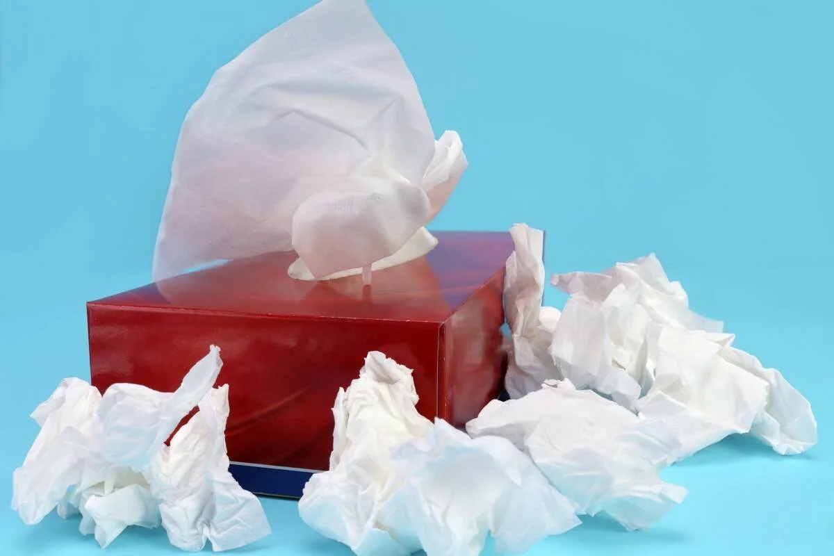 Caja roja de pañuelos de papel rodeada de varios pañuelos arrugados y usados sobre fondo azul claro.