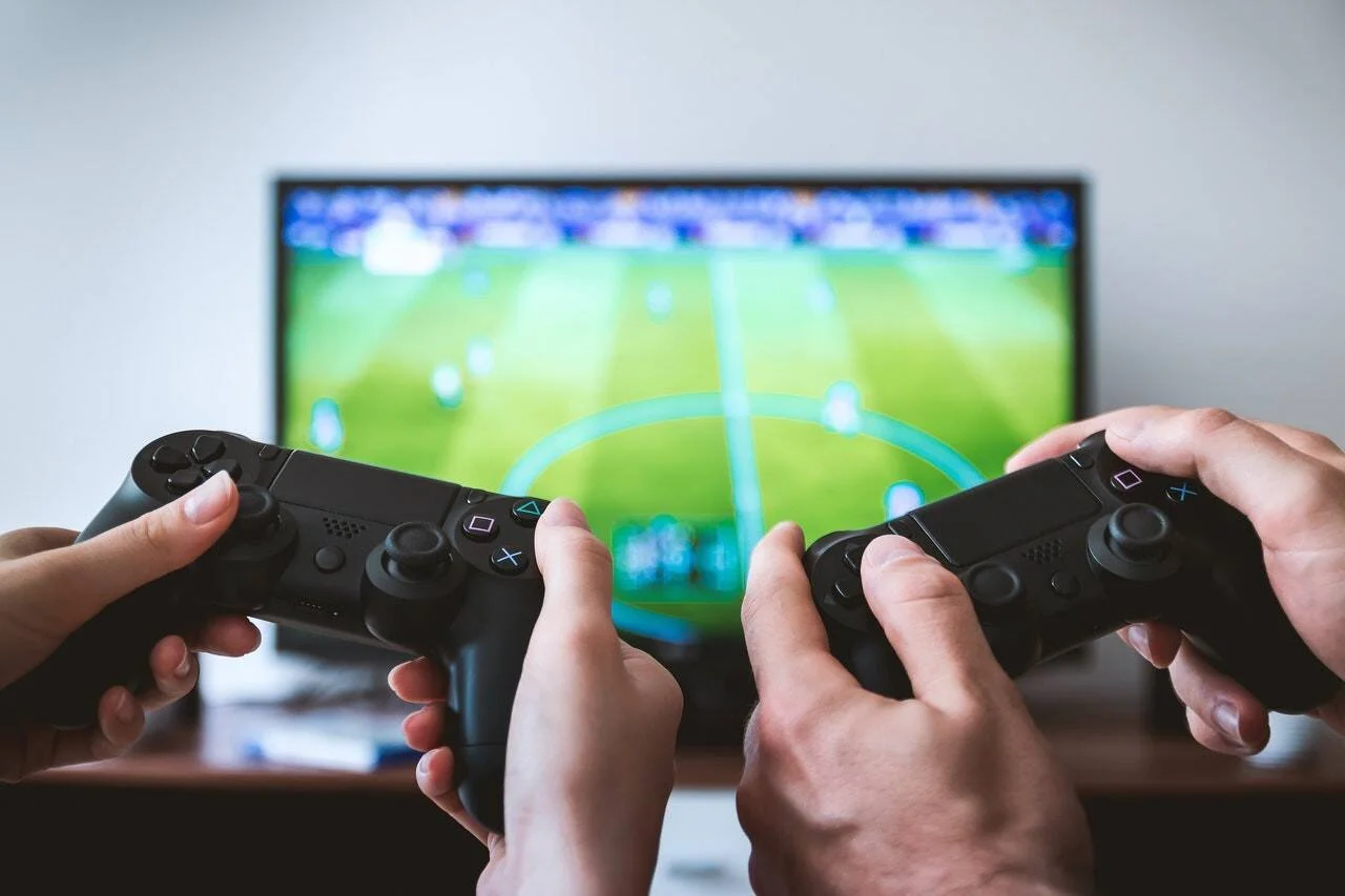 Dos personas sostienen controles de PlayStation frente a una televisión con un videojuego de fútbol en pantalla.
