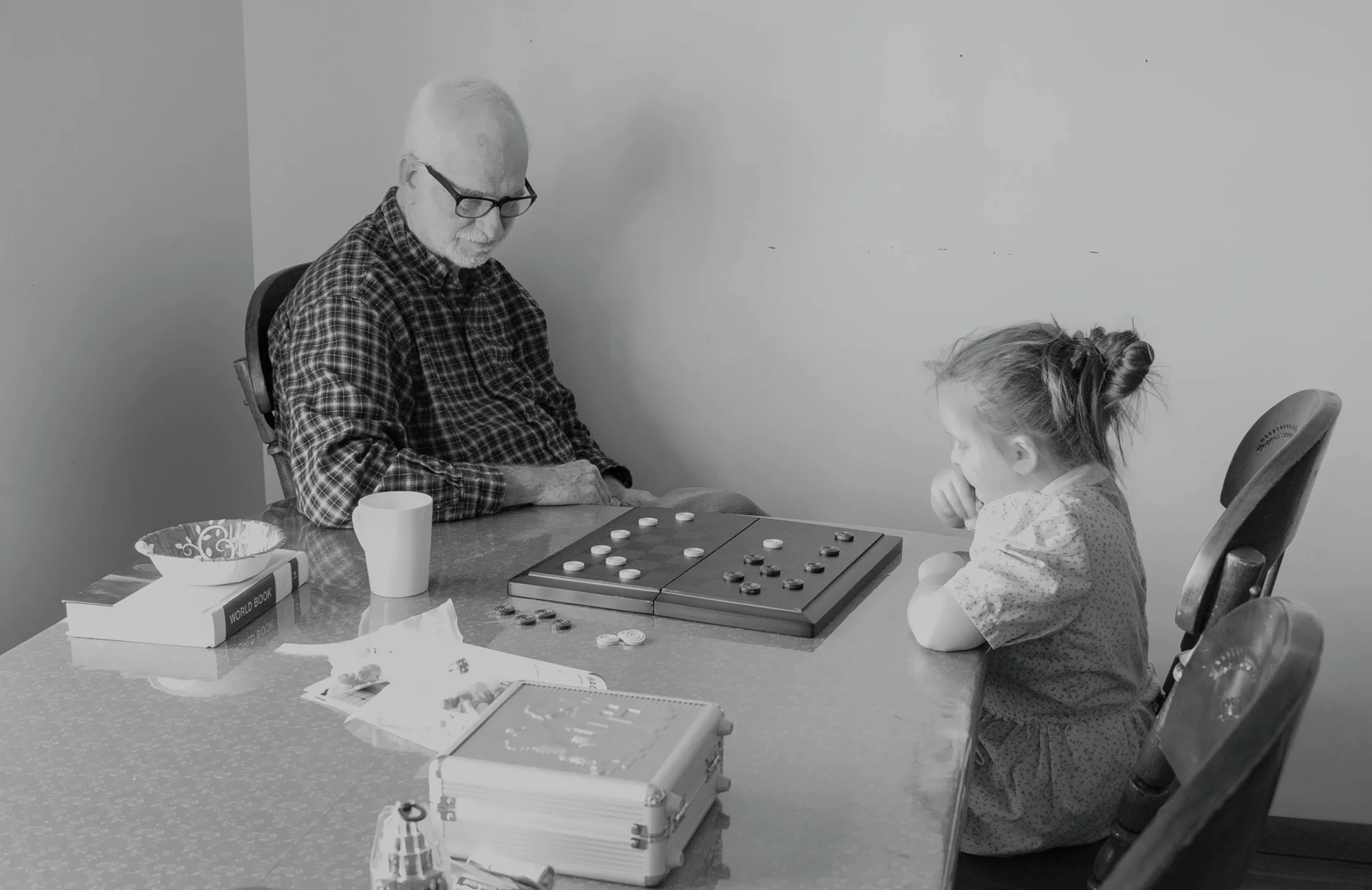 Hombre mayor con lentes y niña pequeña juegan damas sentados frente a frente en una mesa.