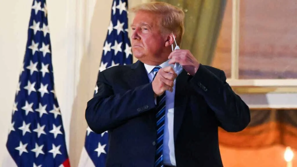 Hombre con traje oscuro y corbata azul se retira una mascarilla frente a banderas de Estados Unidos.