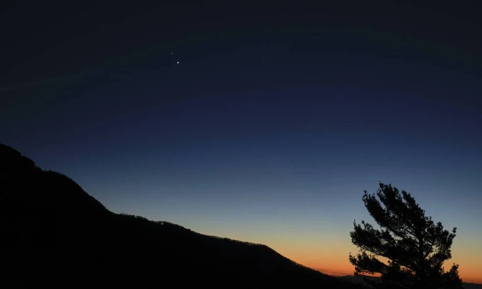 Dos puntos luminosos visibles en el cielo al atardecer sobre la silueta de montañas y un árbol.