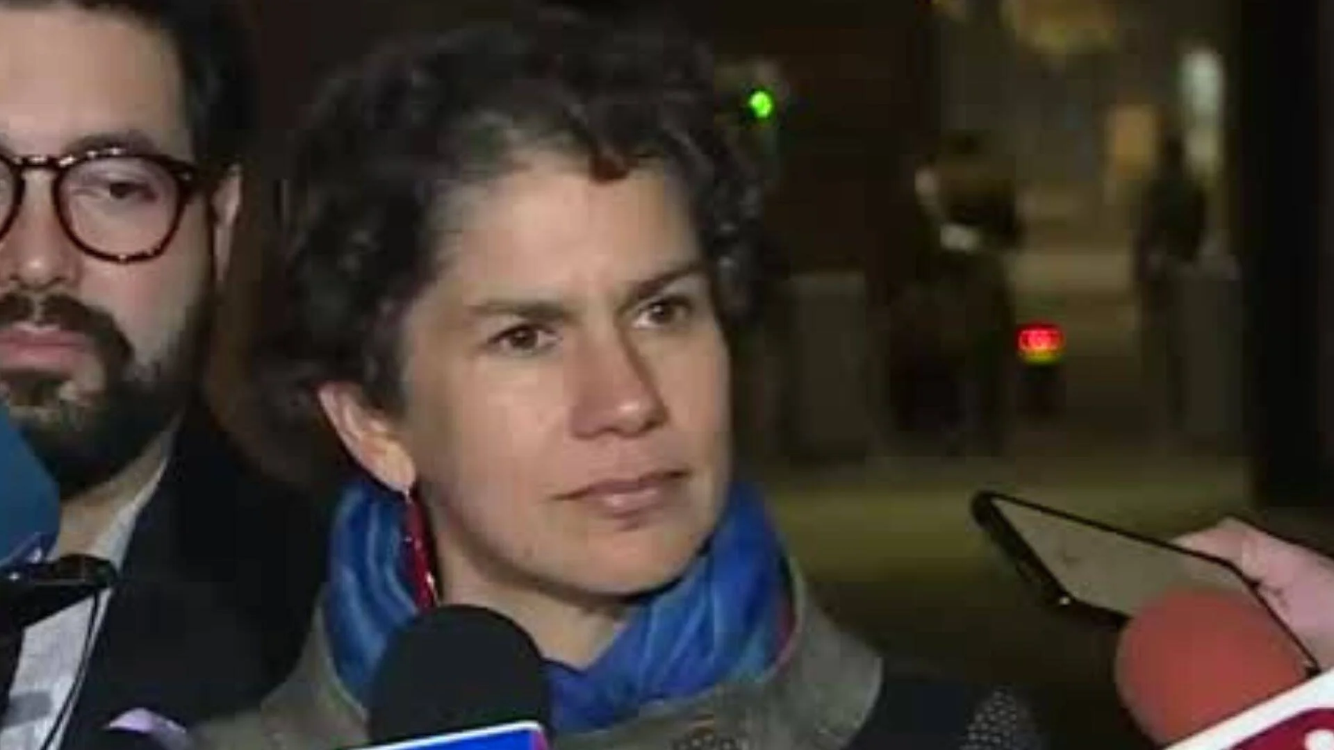Una mujer con pañuelo azul atiende a varios micrófonos de prensa en exteriores nocturnos, acompañada de un hombre con gafas.