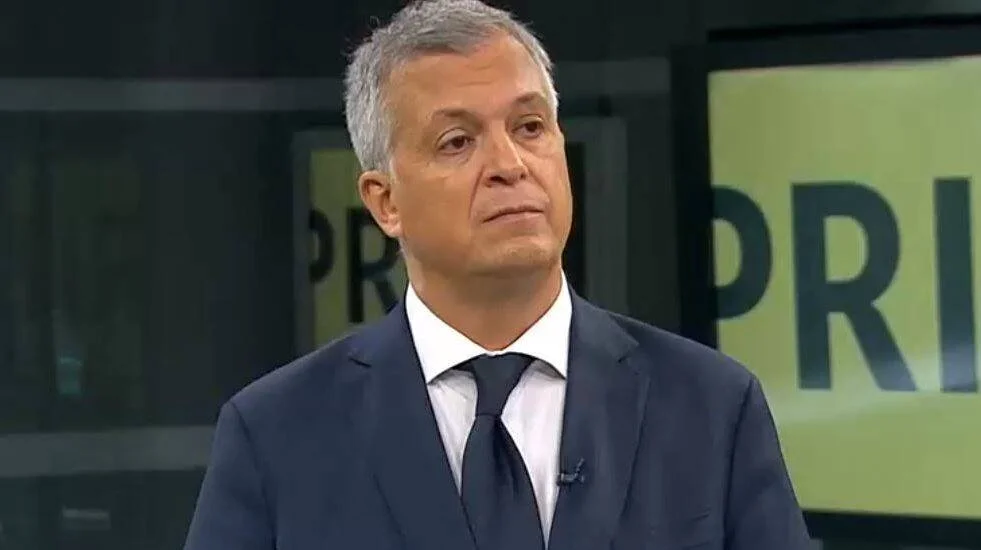 Hombre de cabello canoso con traje azul marino y corbata oscura durante una entrevista en un estudio de televisión.