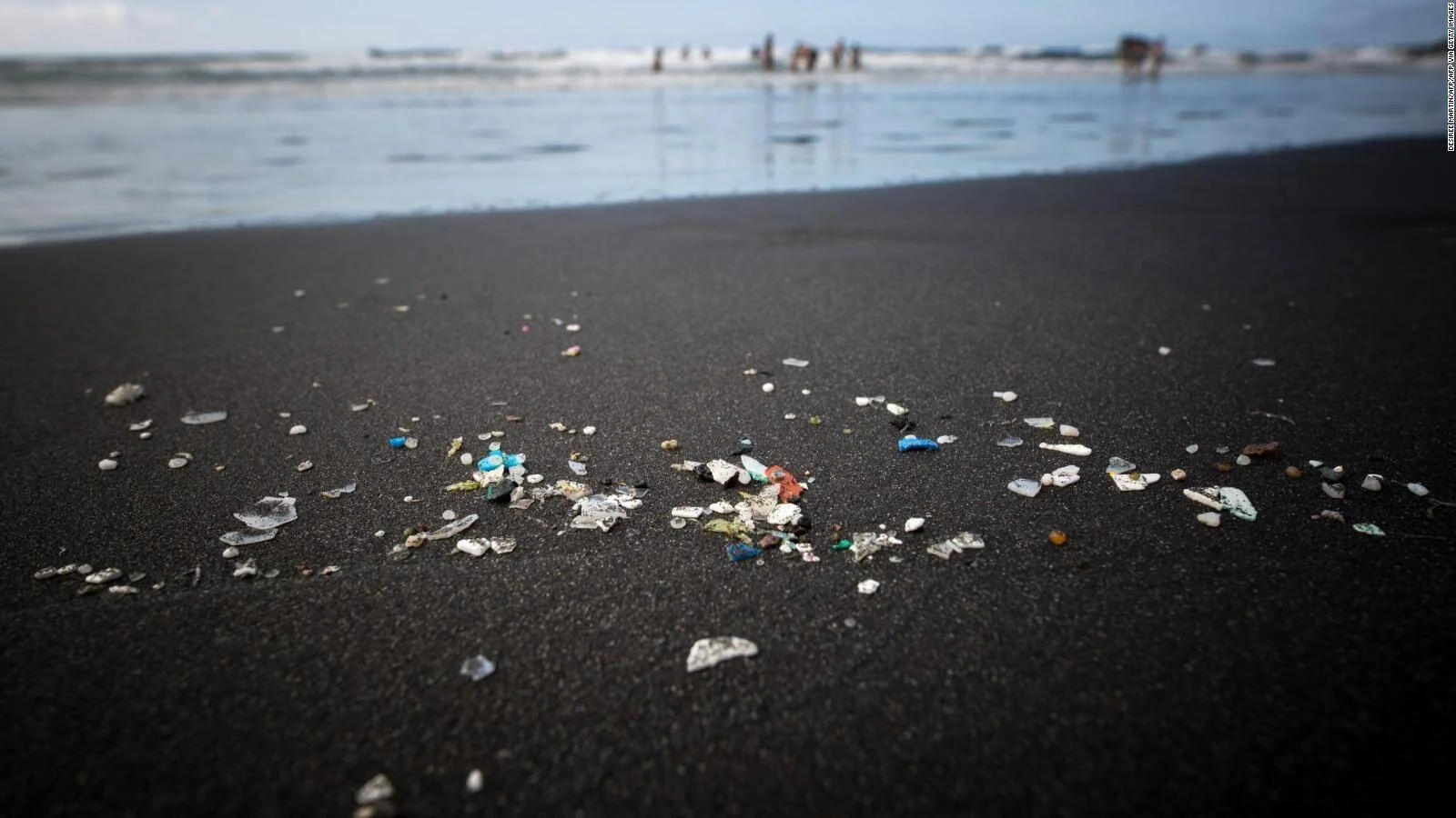 Trozos de plástico y microplásticos dispersos sobre arena oscura de playa, con bañistas al fondo junto al mar.
