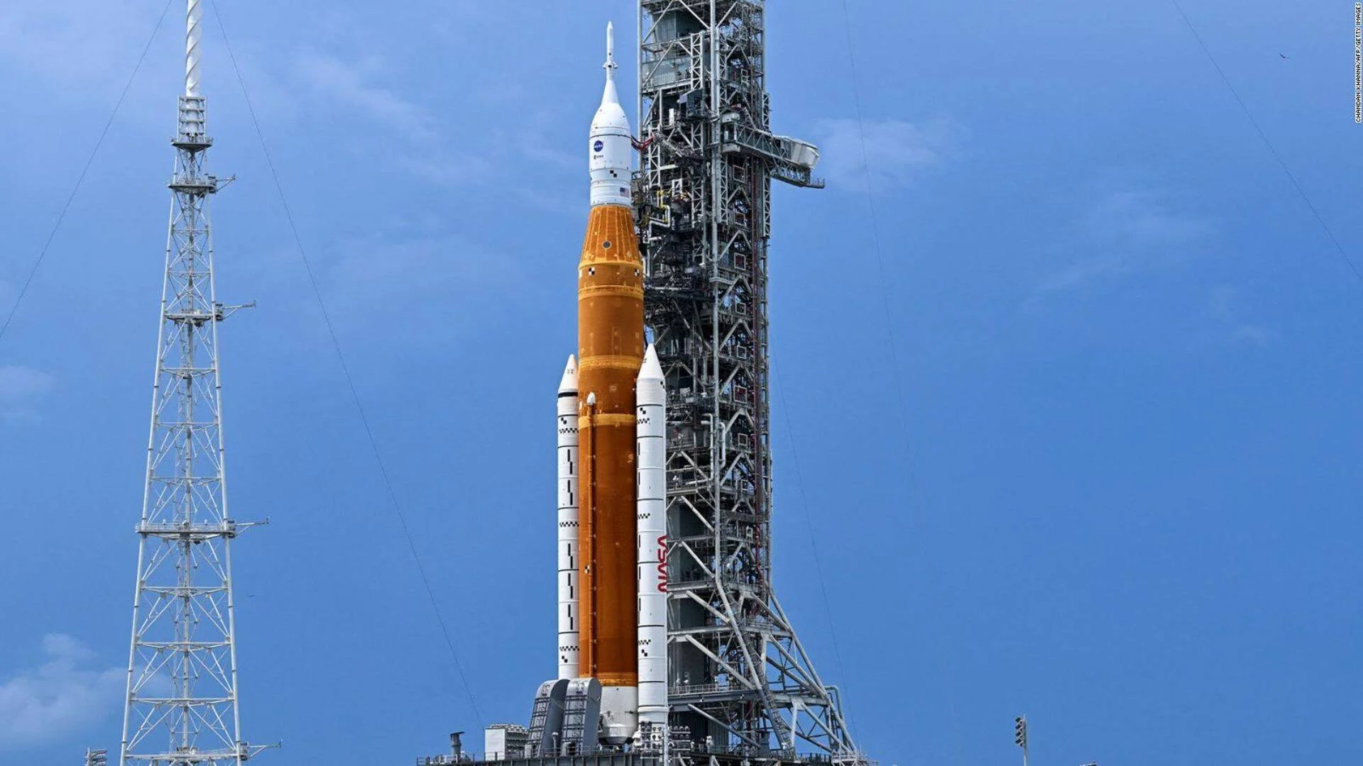 Cohete de la NASA con tanque naranja y propulsores blancos erguido en la torre de lanzamiento bajo cielo azul.
