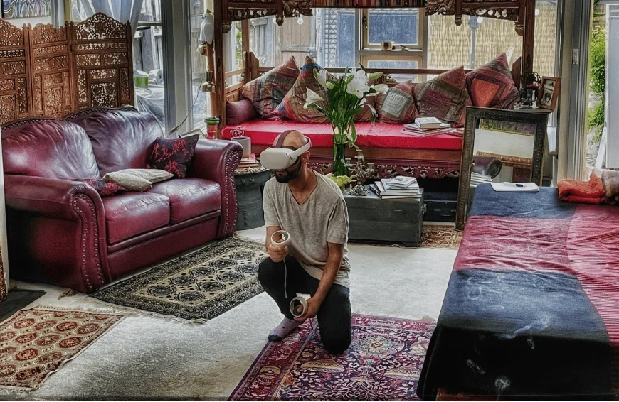 Un hombre arrodillado en el suelo usa un casco de realidad virtual con controles en mano dentro de una sala decorada con alfombras y muebles étnicos.