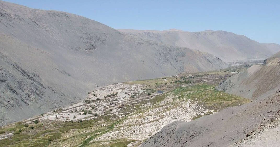 Valle seco flanqueado por cerros áridos con escasa vegetación y pequeñas construcciones dispersas en el fondo.