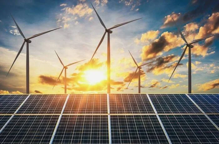 Paneles solares en primer plano y aerogeneradores al fondo bajo un cielo nublado con sol, representando energías renovables.