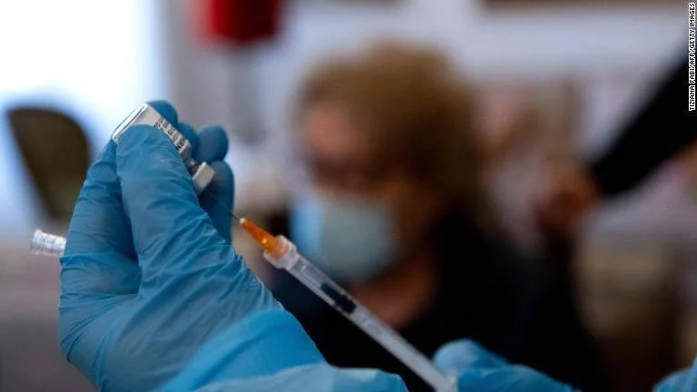 Profesional de salud con guantes azules carga una jeringa con vacuna mientras una persona con mascarilla espera al fondo.