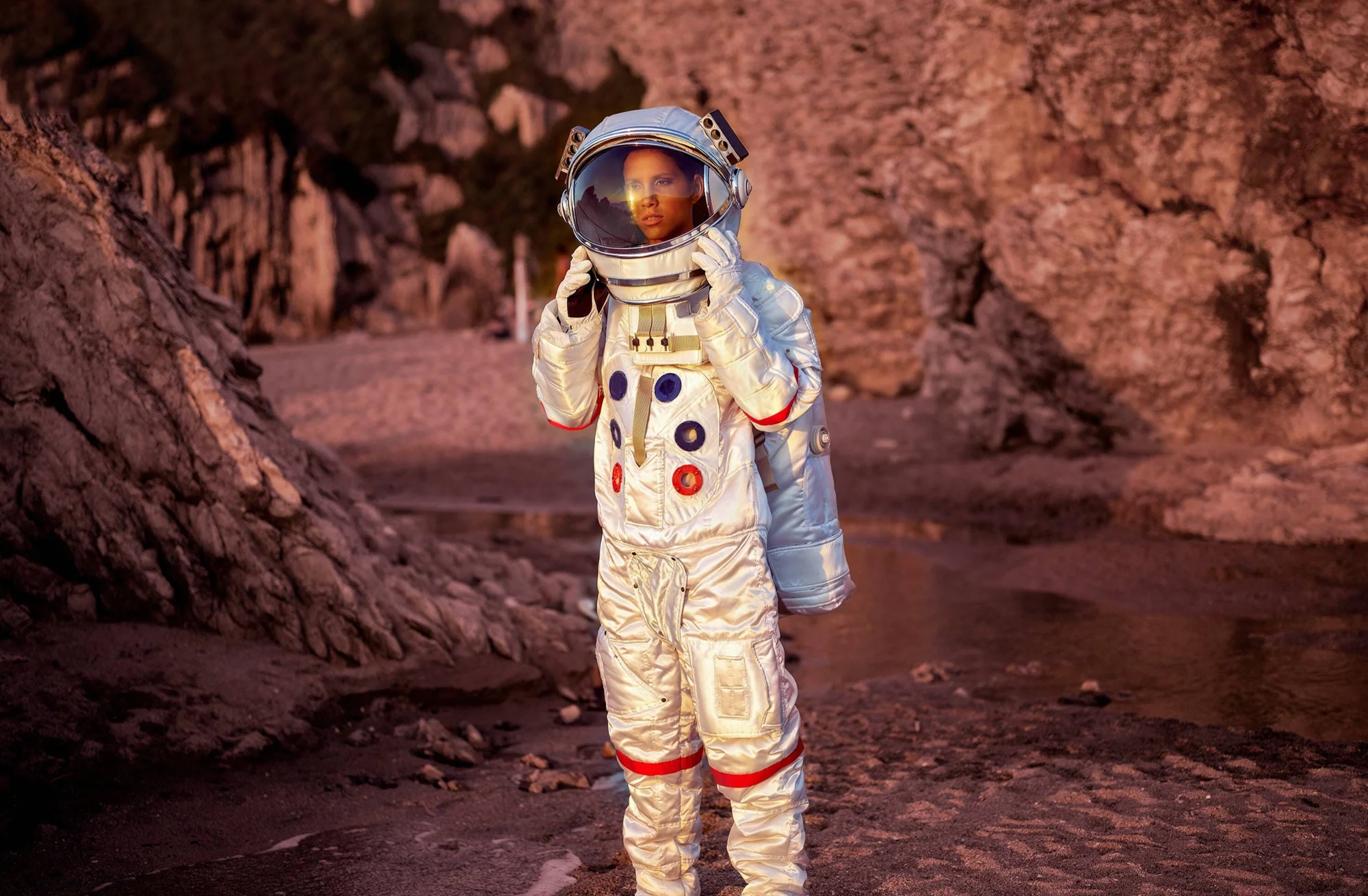 Persona vestida con traje espacial plateado y casco con visor en un terreno rocoso de tonos rojizos, evocando la superficie de un planeta.