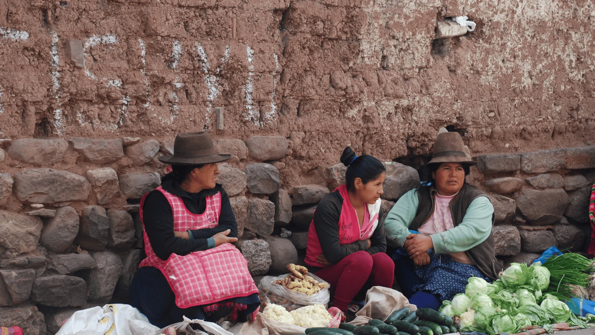 Tres mujeres andinas con delantales rosados y sombreros venden verduras y tubérculos sentadas junto a una pared de piedra.