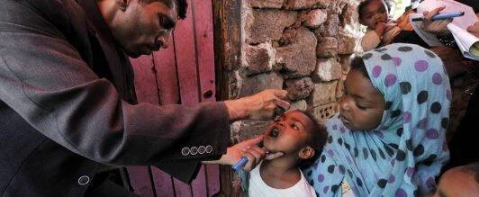 Un trabajador de salud administra la vacuna oral antipolio a un niño pequeño sostenido por una mujer con hiyab.