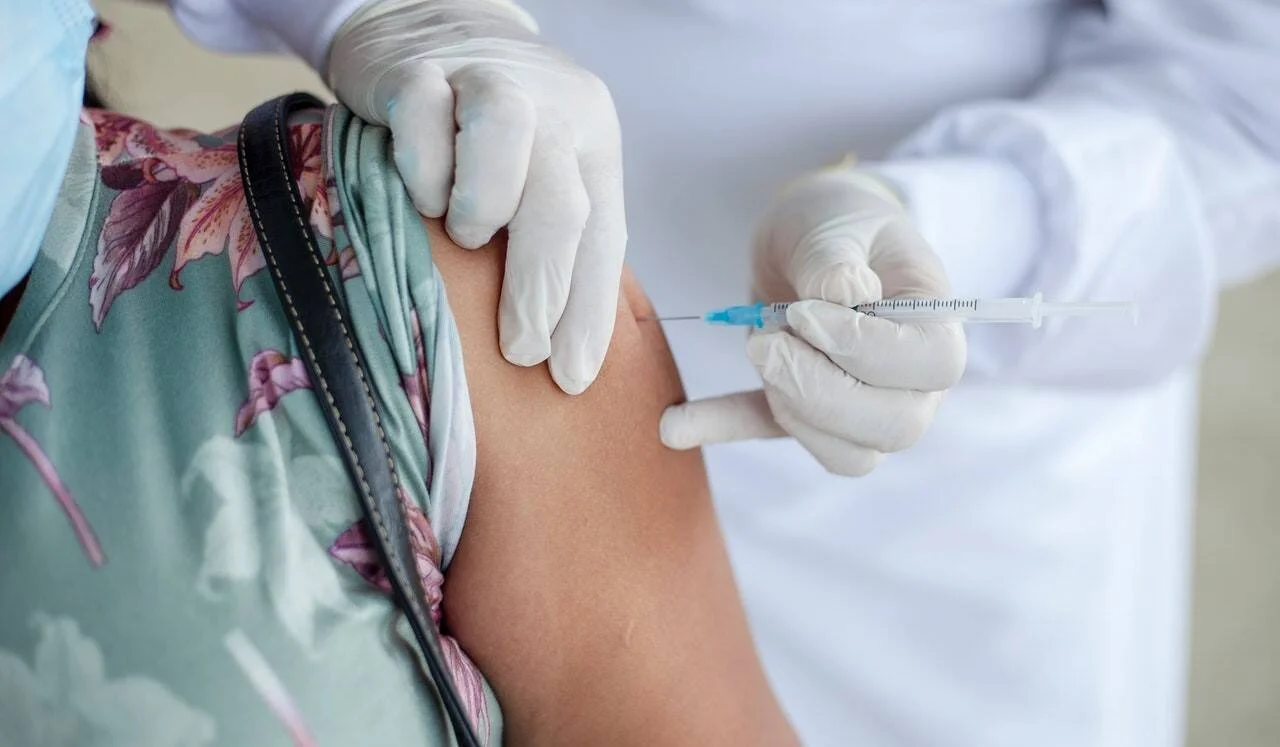 Profesional de salud con guantes aplica una inyección en el brazo de una persona con remera floral.