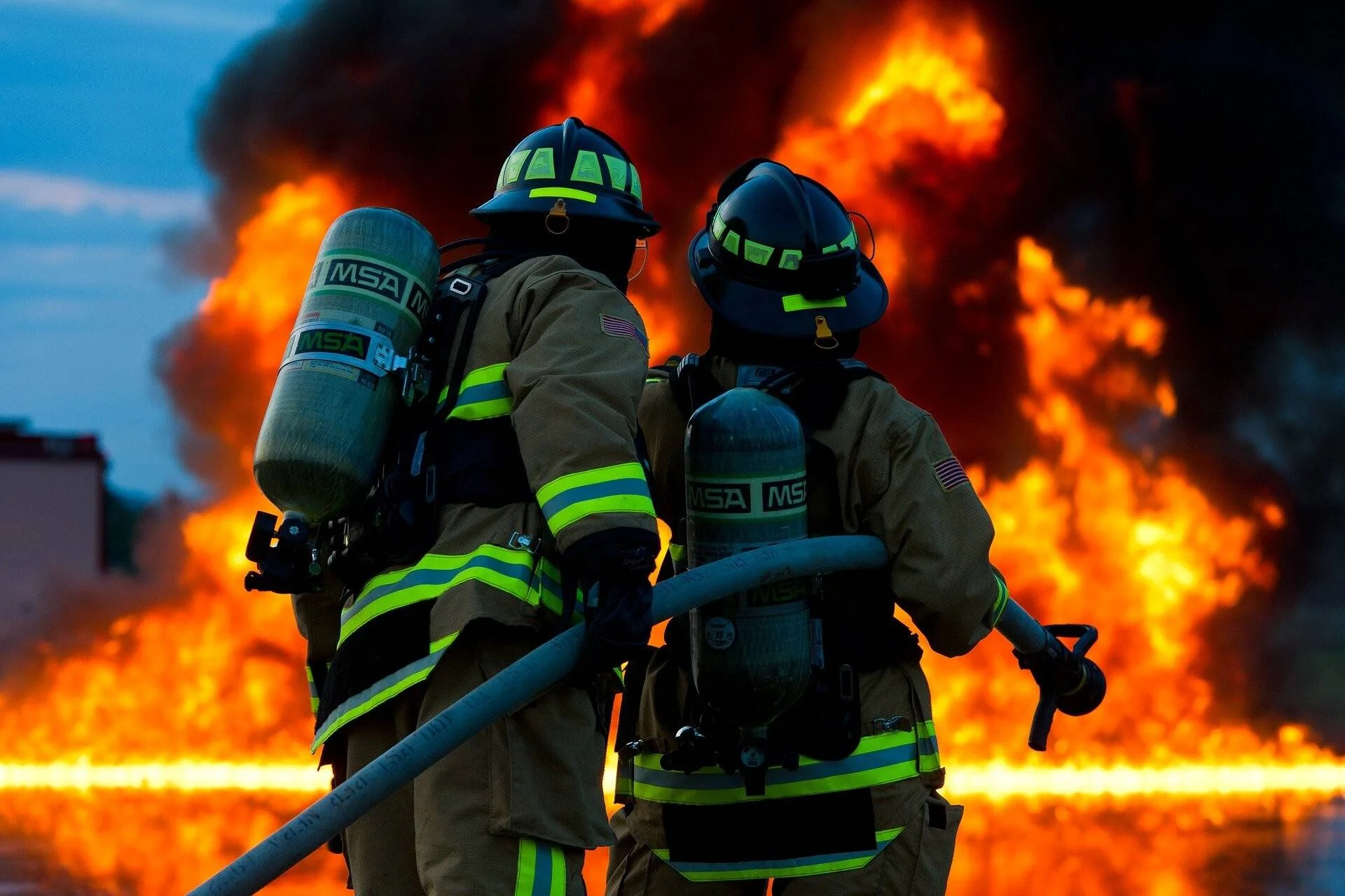 Dos bomberos equipados con trajes ignífugos y tanques de oxígeno MSA sostienen una manguera frente a grandes llamas y humo negro.
