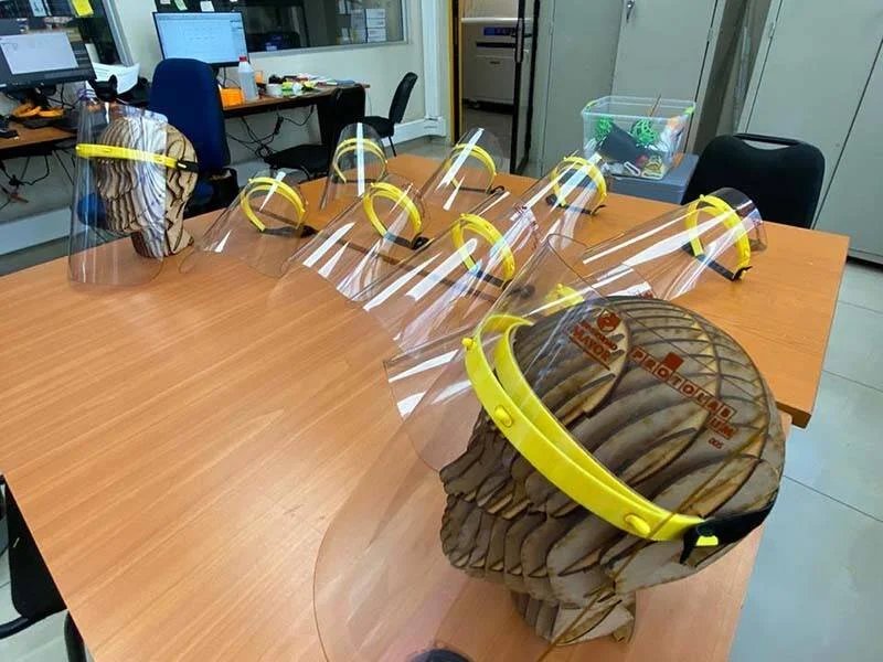 Varias máscaras protectoras faciales con visor transparente y diadema amarilla, apoyadas sobre soportes de cartón en una mesa de trabajo.