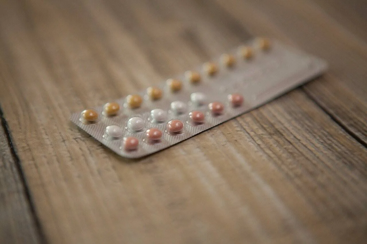 Blíster de píldoras anticonceptivas de tres colores —amarillas, blancas y rosadas— apoyado sobre una superficie de madera.