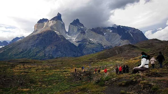Grupo de turistas recorre un sendero con Los Cuernos del Paine al fondo bajo cielo nublado, en la Patagonia chilena.
