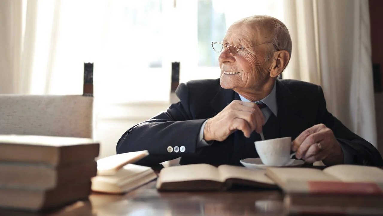Hombre mayor con traje y gafas sonríe mirando hacia un lado, sentado ante una mesa con libros abiertos y una taza de café.