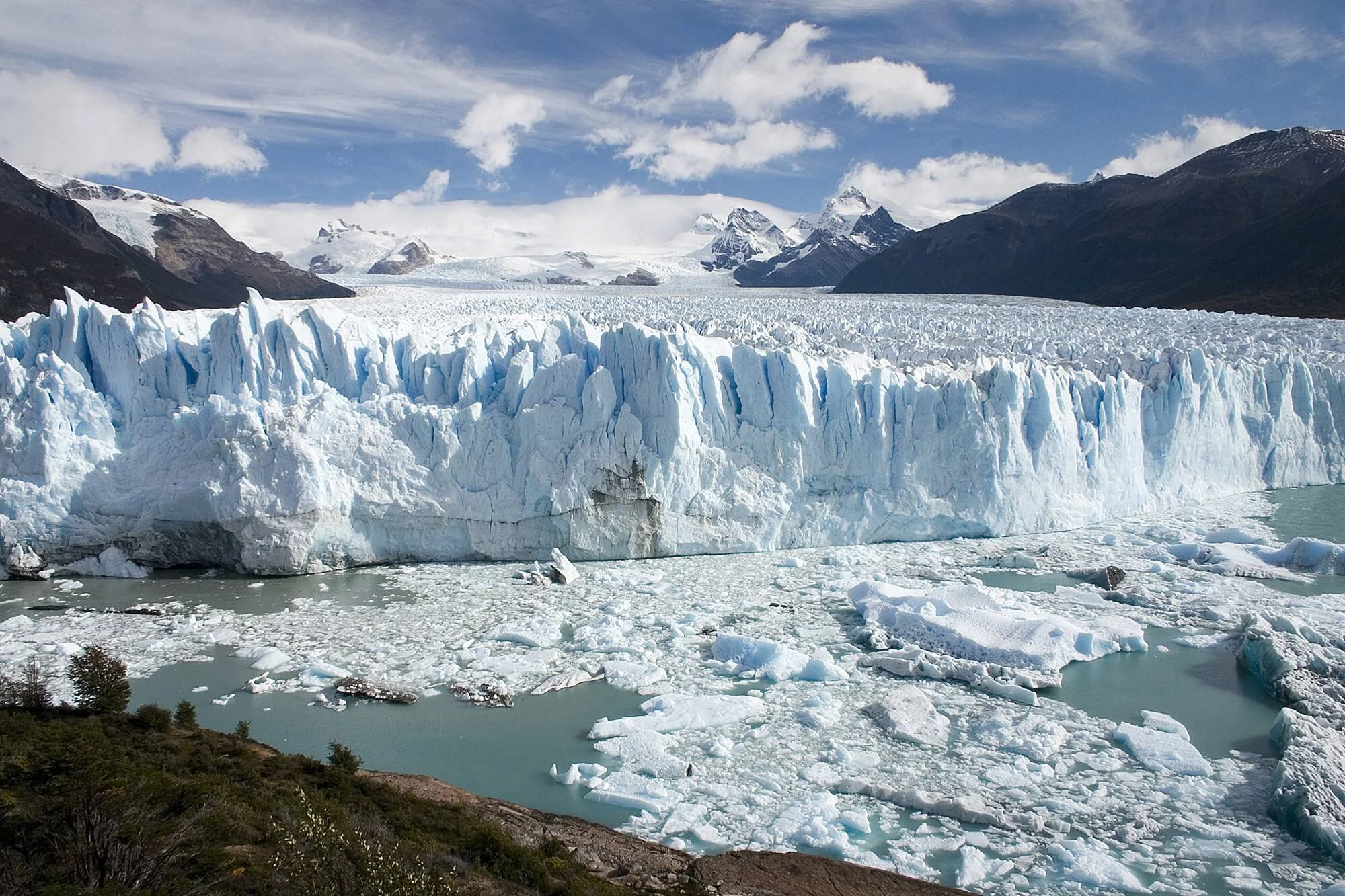 Enorme pared de hielo azul de un glaciar patagónico con témpanos flotando en el lago y montañas nevadas al fondo.