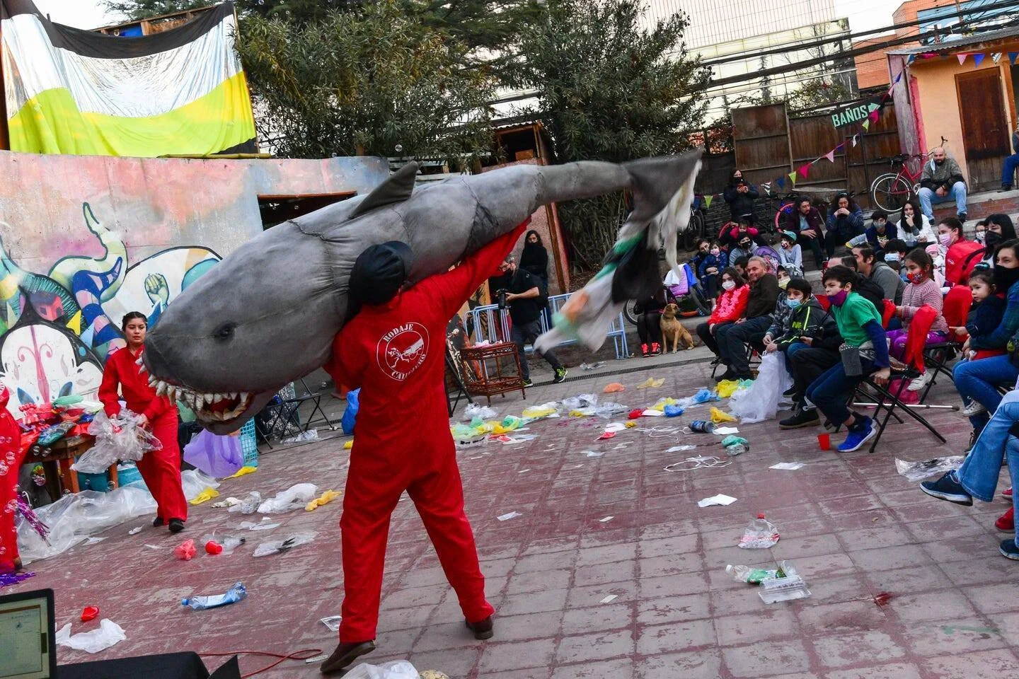 Actor vestido de rojo sostiene en alto una réplica gigante de tiburón ante un público de niños y adultos en una plaza.