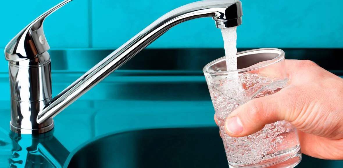 Una mano sostiene un vaso de vidrio que se llena con agua corriente proveniente de un grifo cromado de cocina.