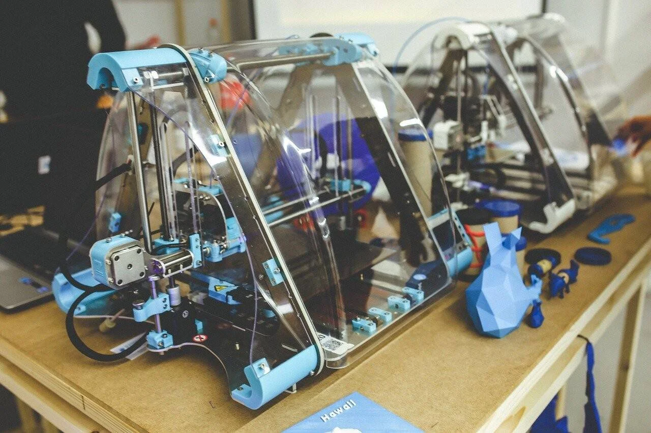 Dos impresoras 3D con estructura azul y cubierta acrílica transparente sobre una mesa, junto a objetos impresos en plástico azul.