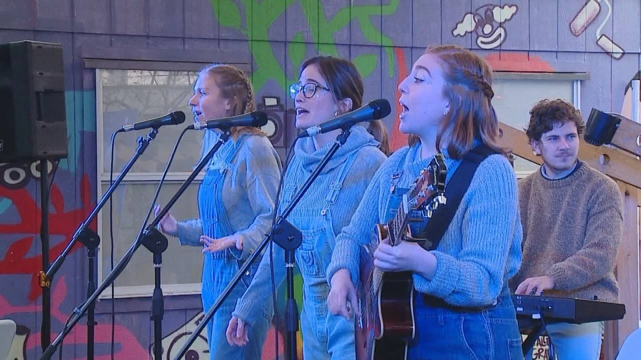 Tres jóvenes cantan ante micrófonos en un escenario con murales, una toca guitarra y un músico al fondo en teclado.