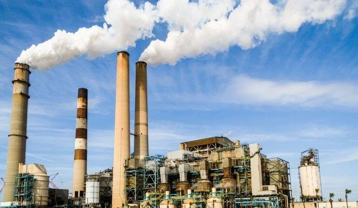 Complejo industrial con cuatro chimeneas expulsando grandes columnas de humo blanco hacia el cielo azul.