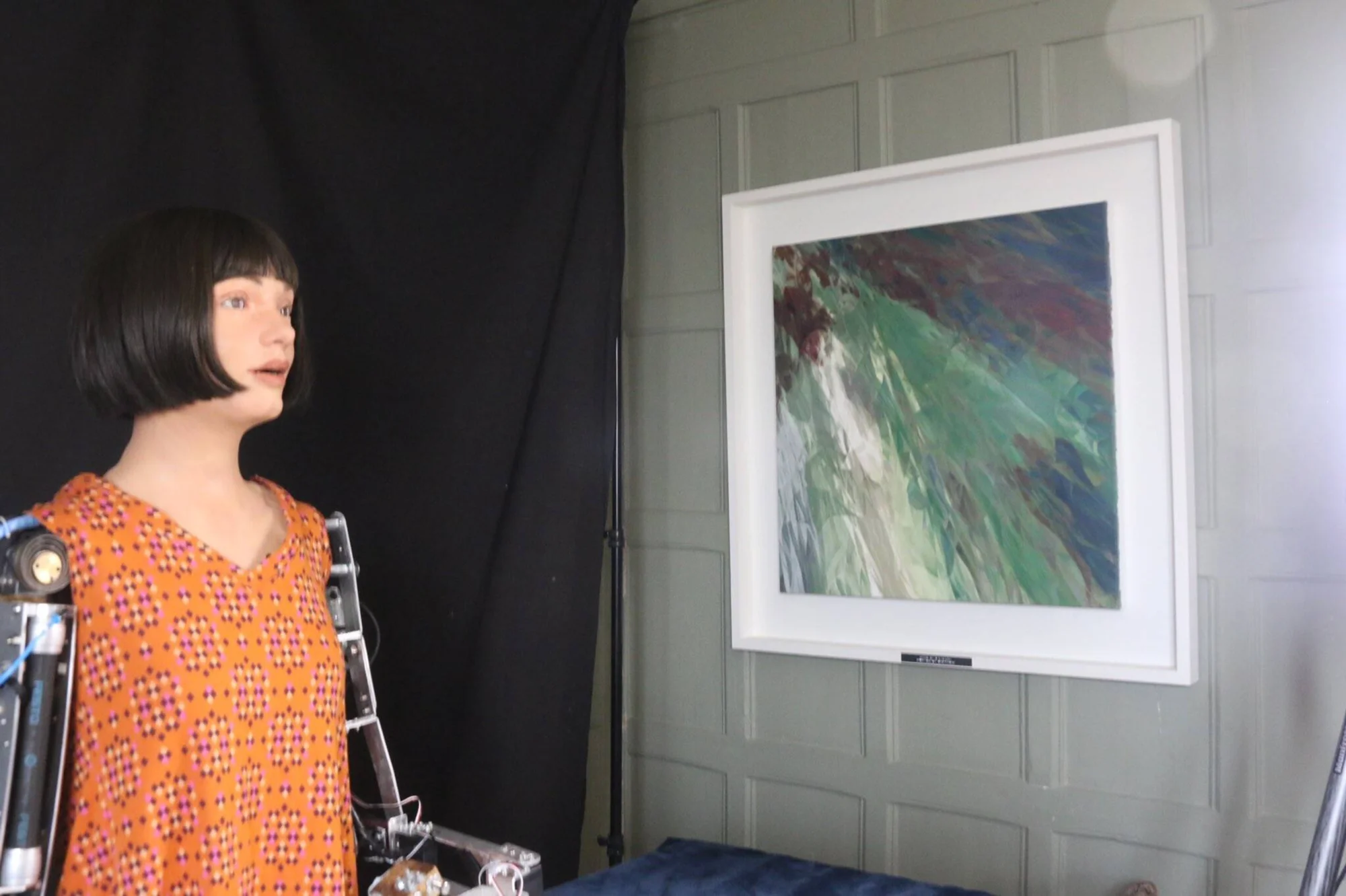 Robot humanoide de apariencia femenina con vestido naranja contempla una pintura abstracta de colores verdes y rojos colgada en la pared.