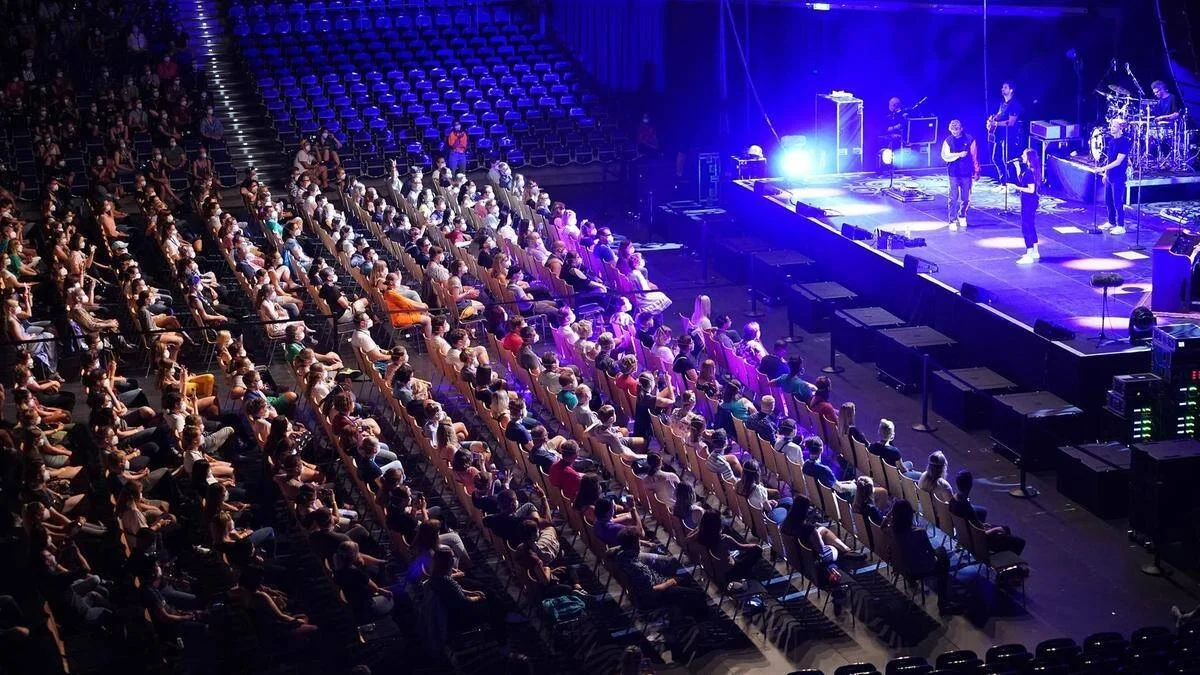 Vista aérea de una sala de conciertos con aforo reducido y butacas vacías mientras una banda actúa en el escenario iluminado.