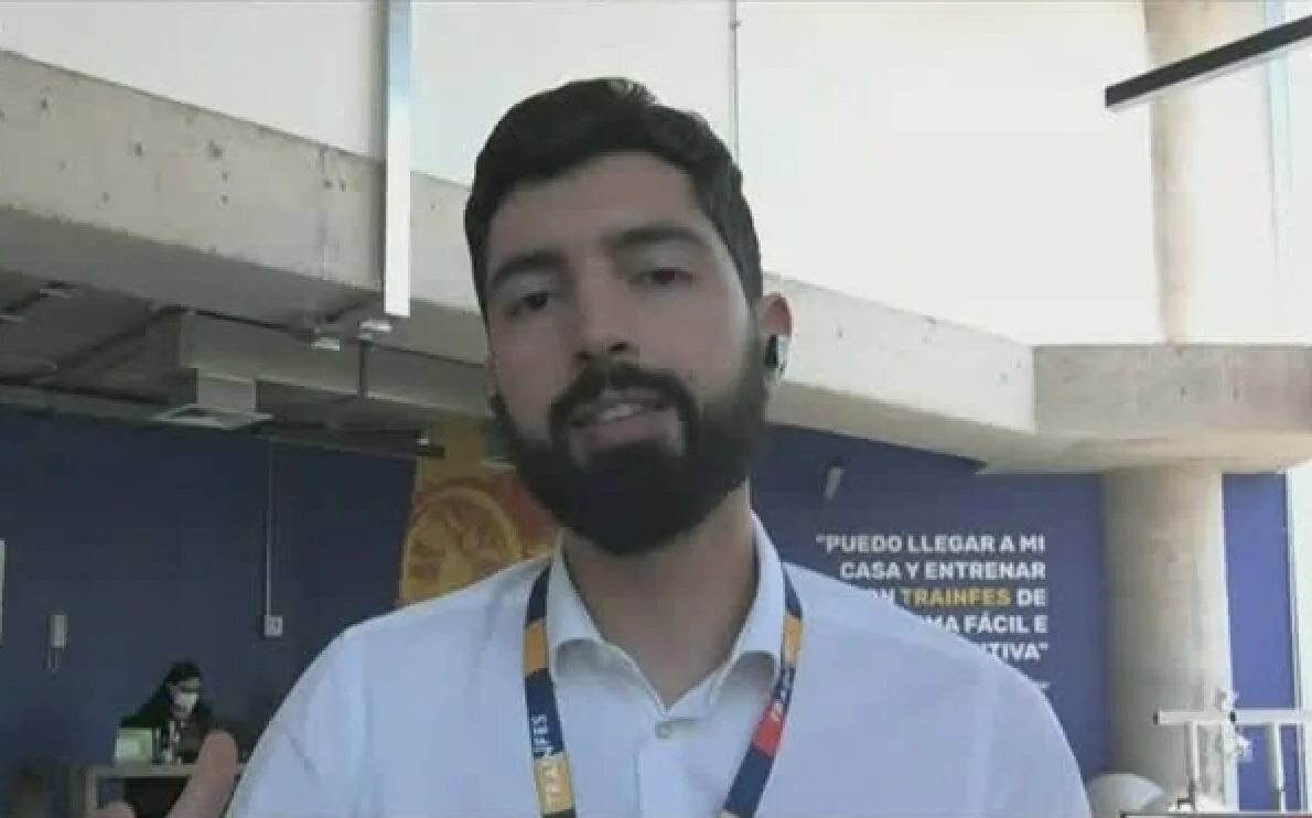Hombre joven con barba y credencial de Trainfes habla frente a un cartel con la frase sobre entrenar en casa.